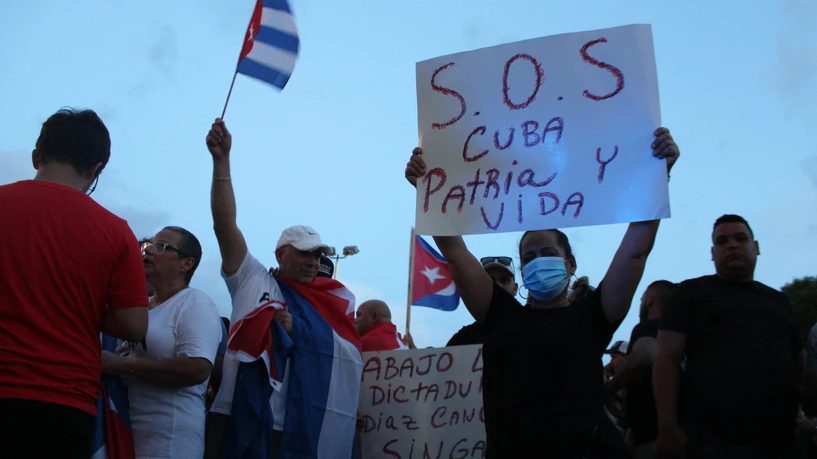 Una mujer con mascarilla sostiene un cartel escrito a mano que dice «S.O.S. Cuba, Patria y Vida»; alrededor, personas con banderas cubanas.