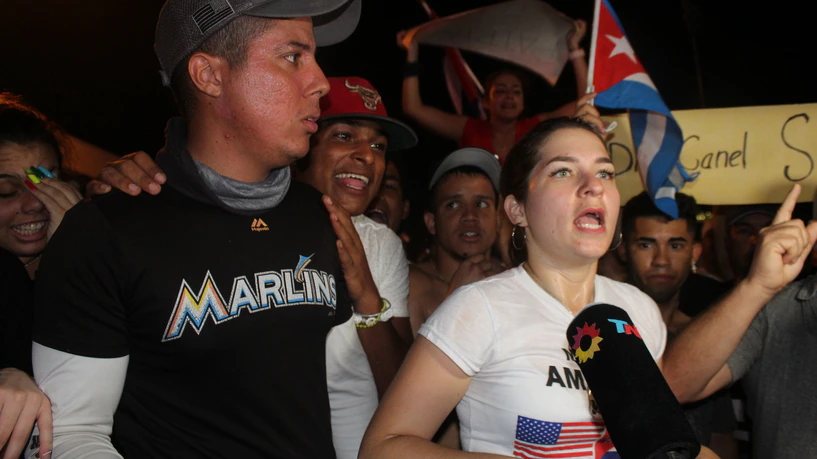 Un grupo de jóvenes grita en una concentración nocturna; una mujer habla ante un micrófono de televisión y detrás ondean banderas cubanas.