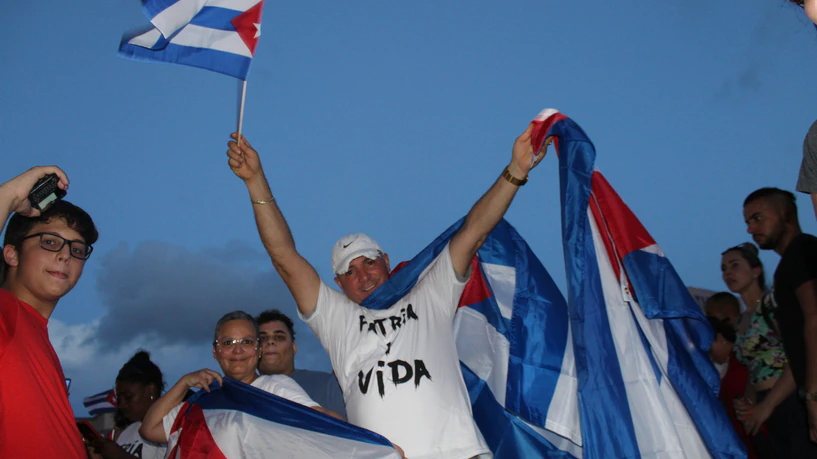 Un hombre con camiseta blanca en la que se lee «Patria y Vida» levanta los brazos con una bandera cubana; al fondo, más banderas.