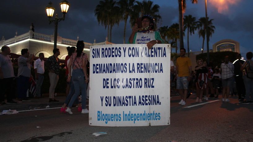 Un hombre sostiene un gran cartel impreso que pide la renuncia de los Castro; detrás, gente en la calle al anochecer.