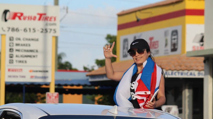 Una joven con gorra y una bandera cubana sobre los hombros, asomada por el techo de un coche, hace con la mano la señal de la L.