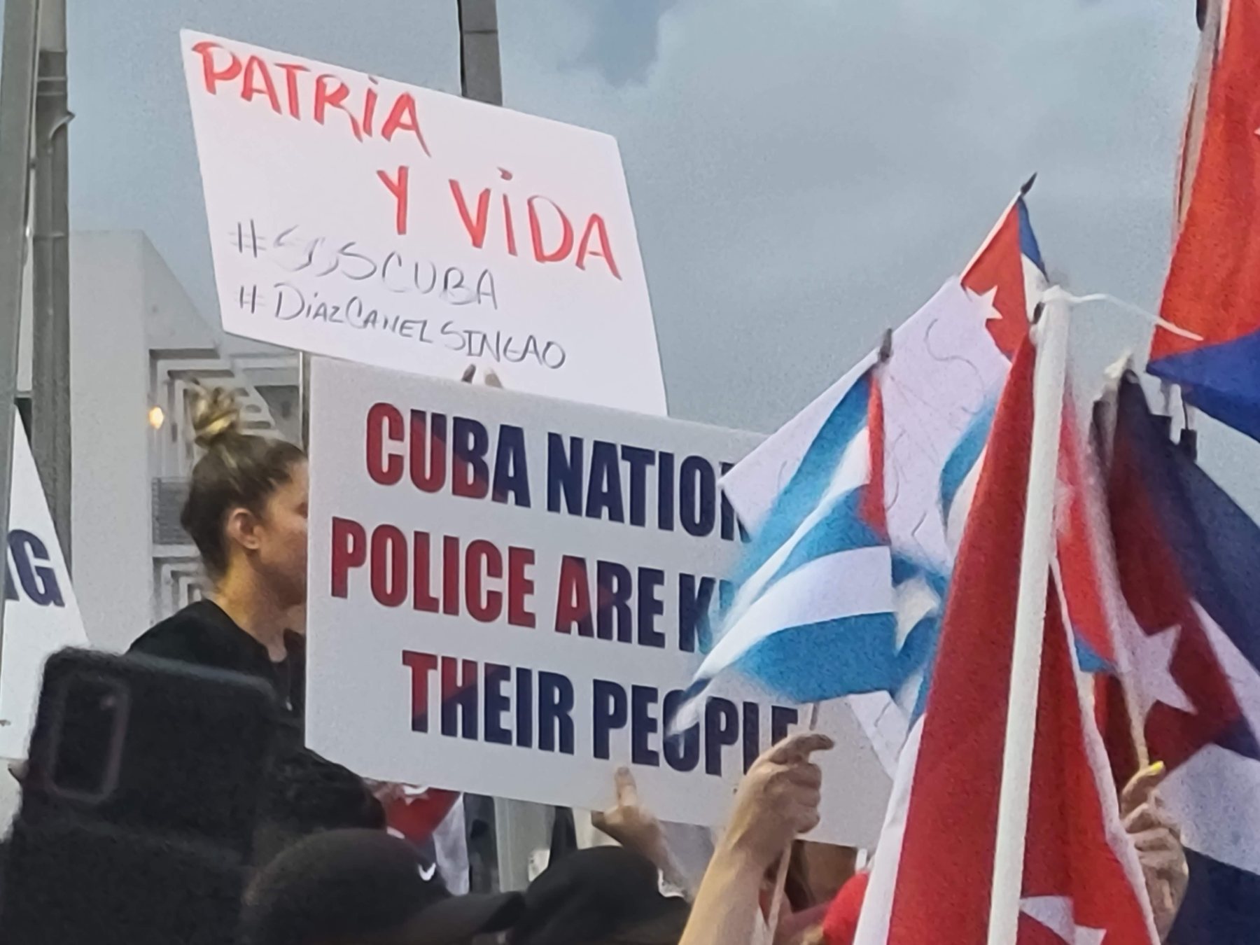 Carteles escritos a mano con «Patria y Vida» y «#SOSCuba» entre banderas cubanas, en una concentración al aire libre.