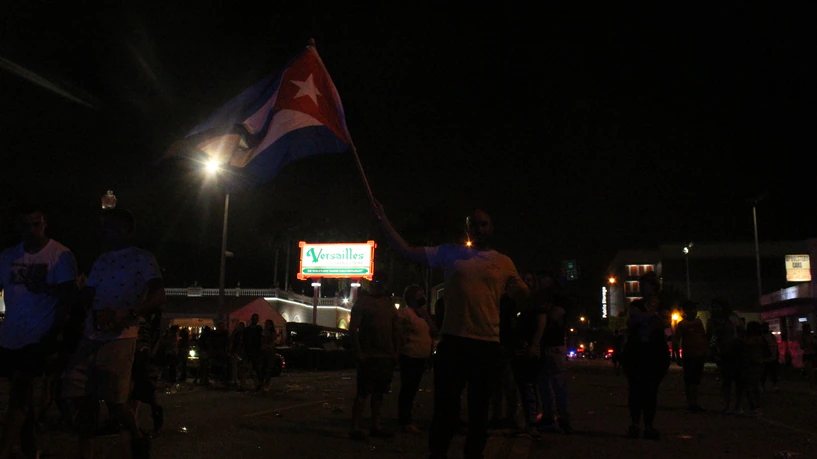 Un hombre ondea una bandera cubana de noche frente al rótulo del restaurante Versailles, con gente alrededor.