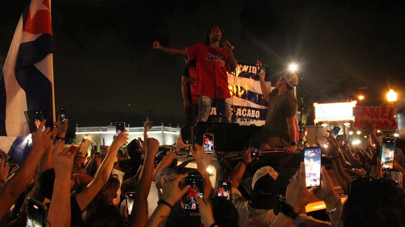 Un cantante con camiseta roja, subido a un vehículo, canta ante una multitud que levanta teléfonos y banderas de noche.