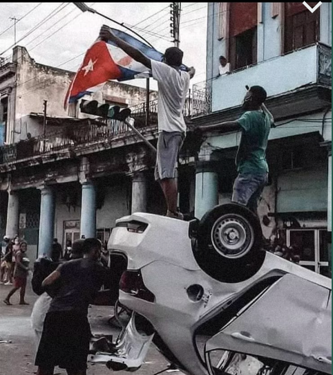 Dos personas sobre un vehículo volcado; una sostiene una bandera cubana.