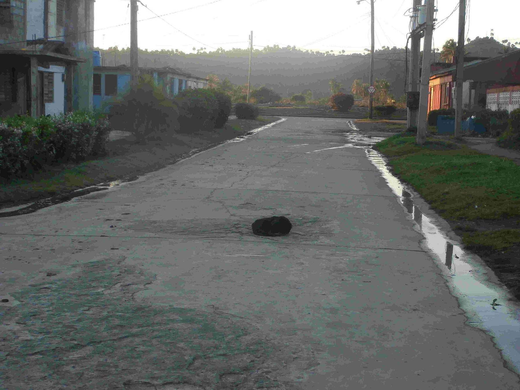 Una calle de hormigón al amanecer con agua corriendo por ambos bordes; casas bajas a los lados y colinas al fondo.
