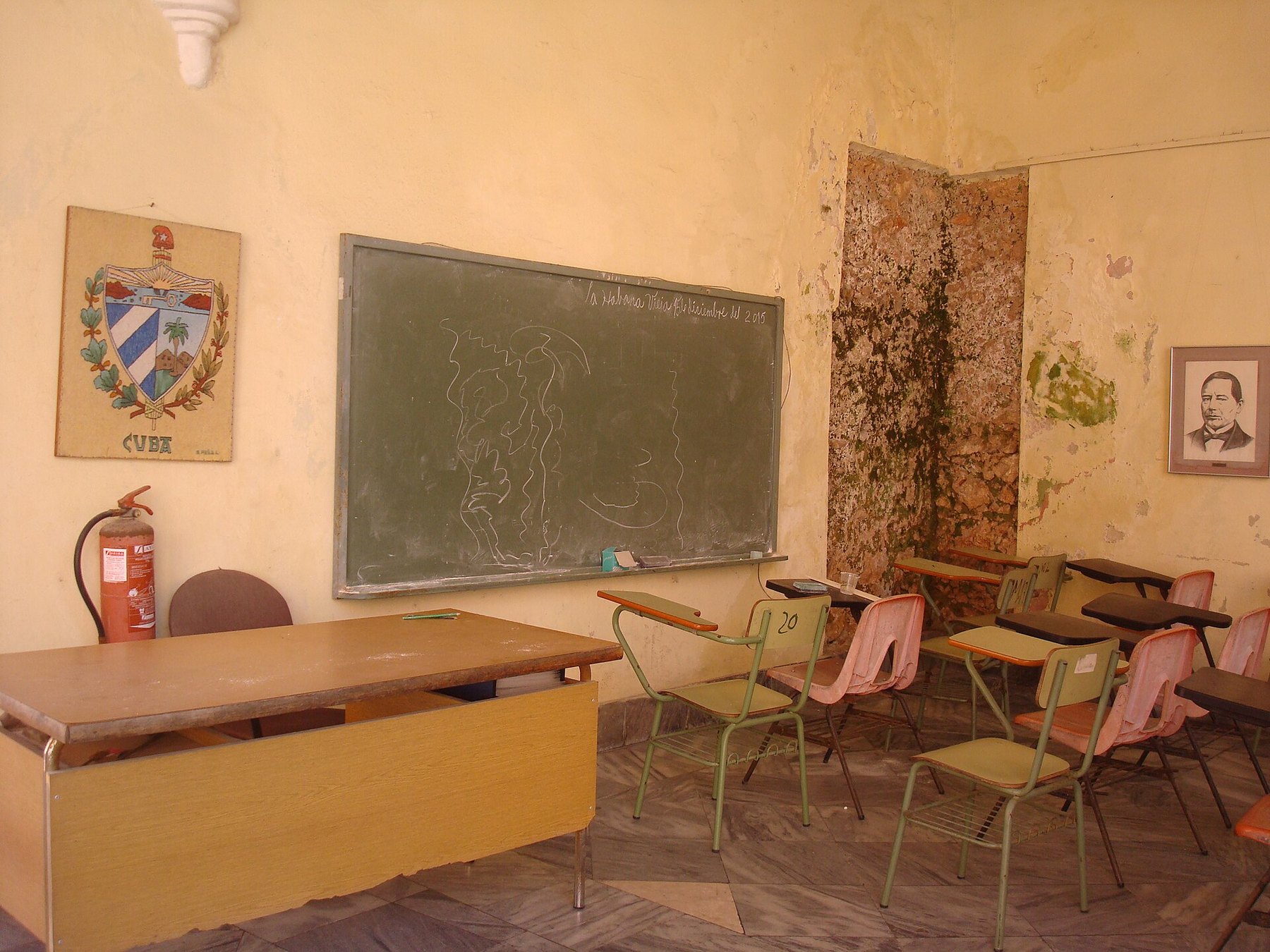 Aula vacía con pizarra verde, un escudo de Cuba pintado en la pared, un extintor, pupitres de metal y una pared con el revoque caído.