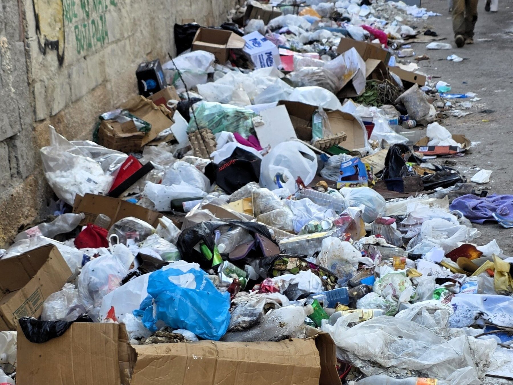 Una acera de La Habana cubierta por bolsas de basura, cartones y botellas a lo largo de un muro; al fondo caminan dos personas.