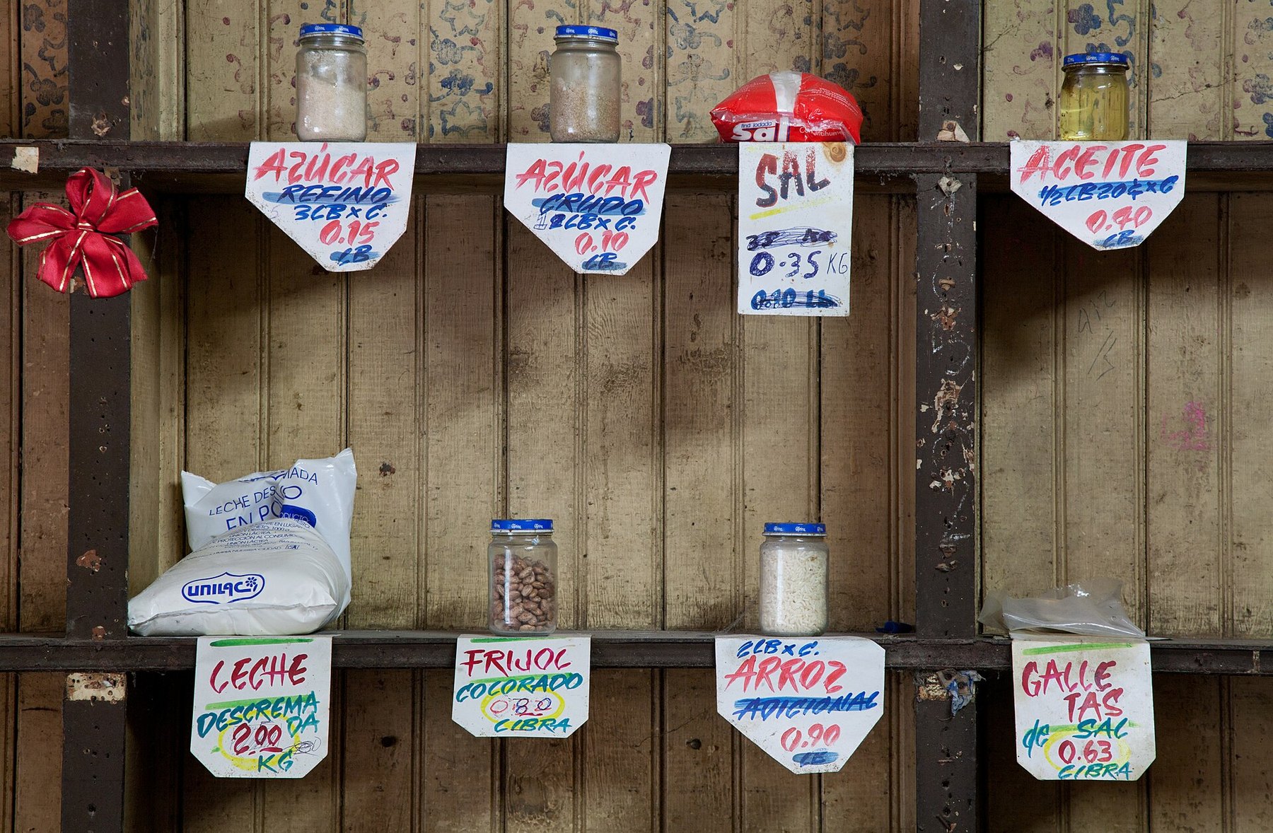 Estante de madera de una bodega con frascos de azúcar, sal, aceite, frijoles y arroz, y carteles escritos a mano con los precios en centavos.