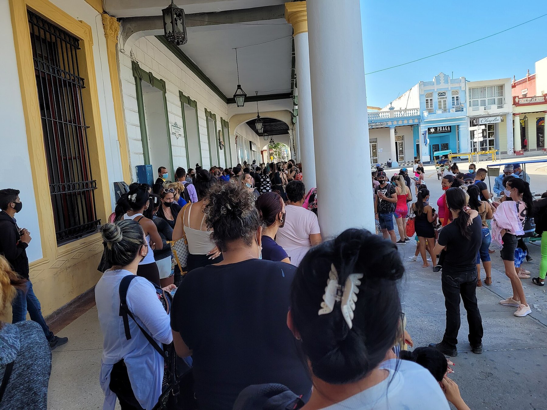 Decenas de personas, muchas con mascarilla, hacen cola bajo los soportales de un edificio colonial en una plaza soleada.