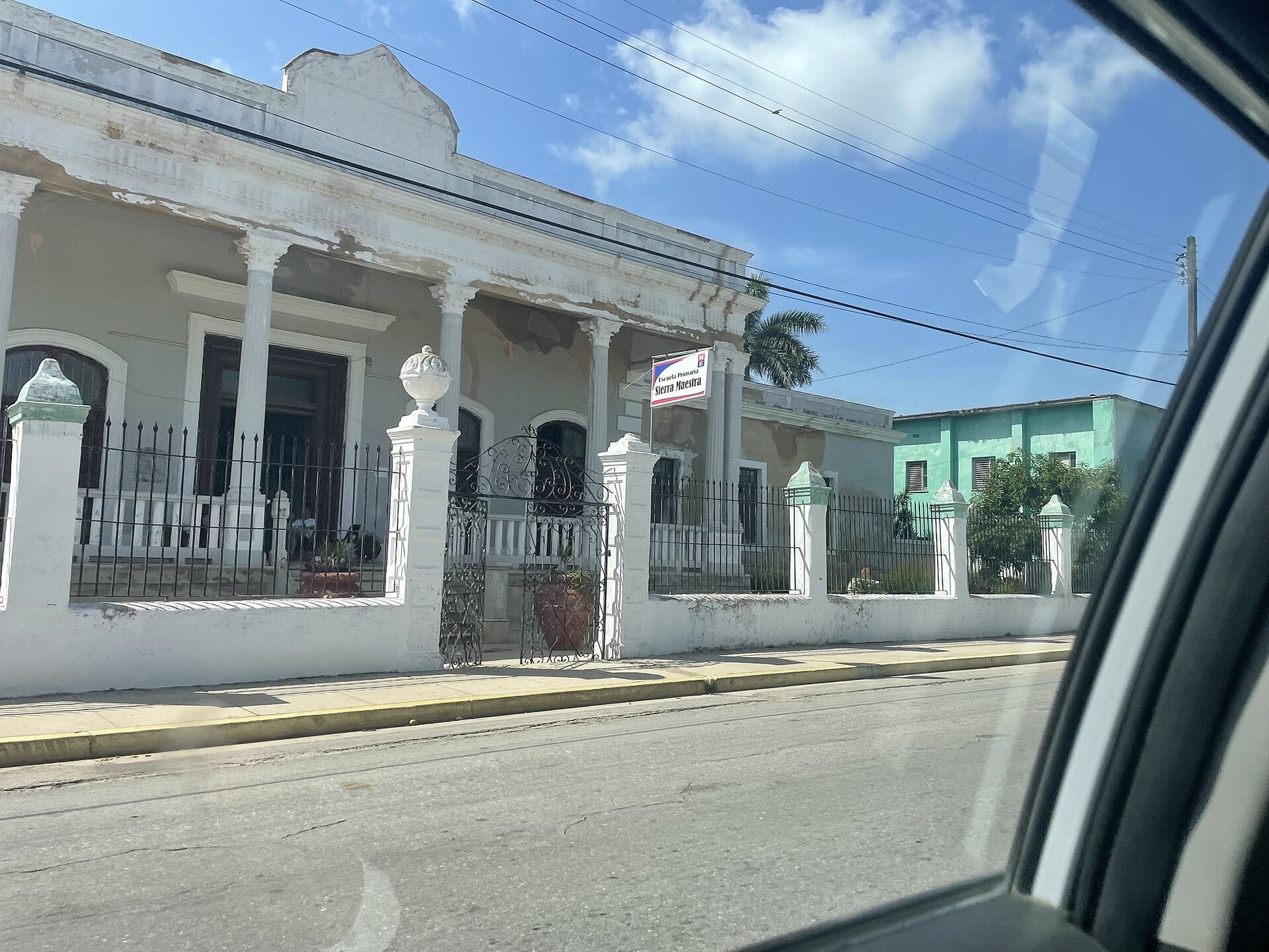Fachada de una casa colonial blanca con columnas y verja de hierro, con un cartel de escuela primaria, vista desde la ventanilla de un coche.