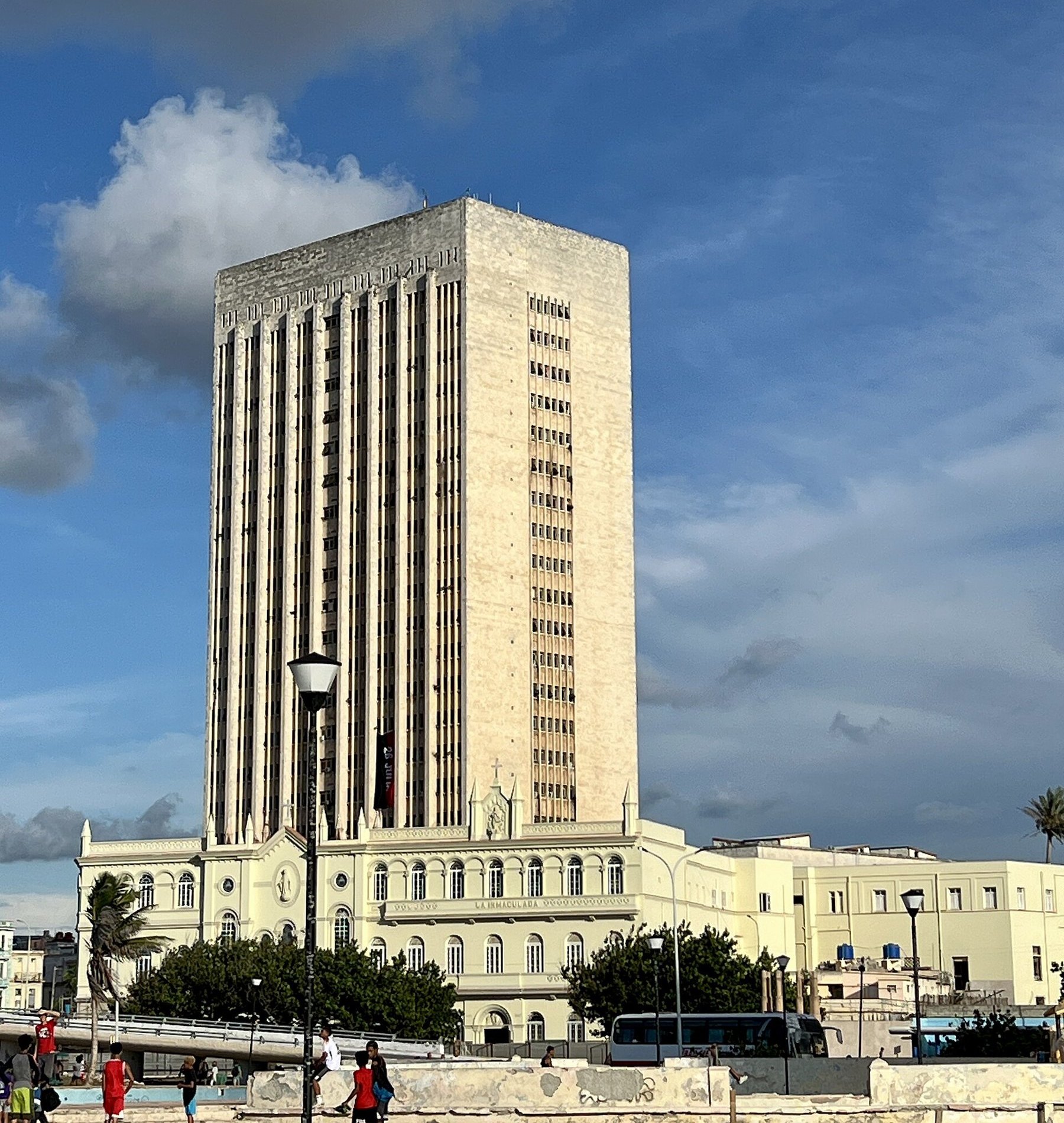 Torre de hormigón claro de veinticuatro plantas con ventanas estrechas, sobre un edificio antiguo de color crema; delante, un muro con niños jugando.