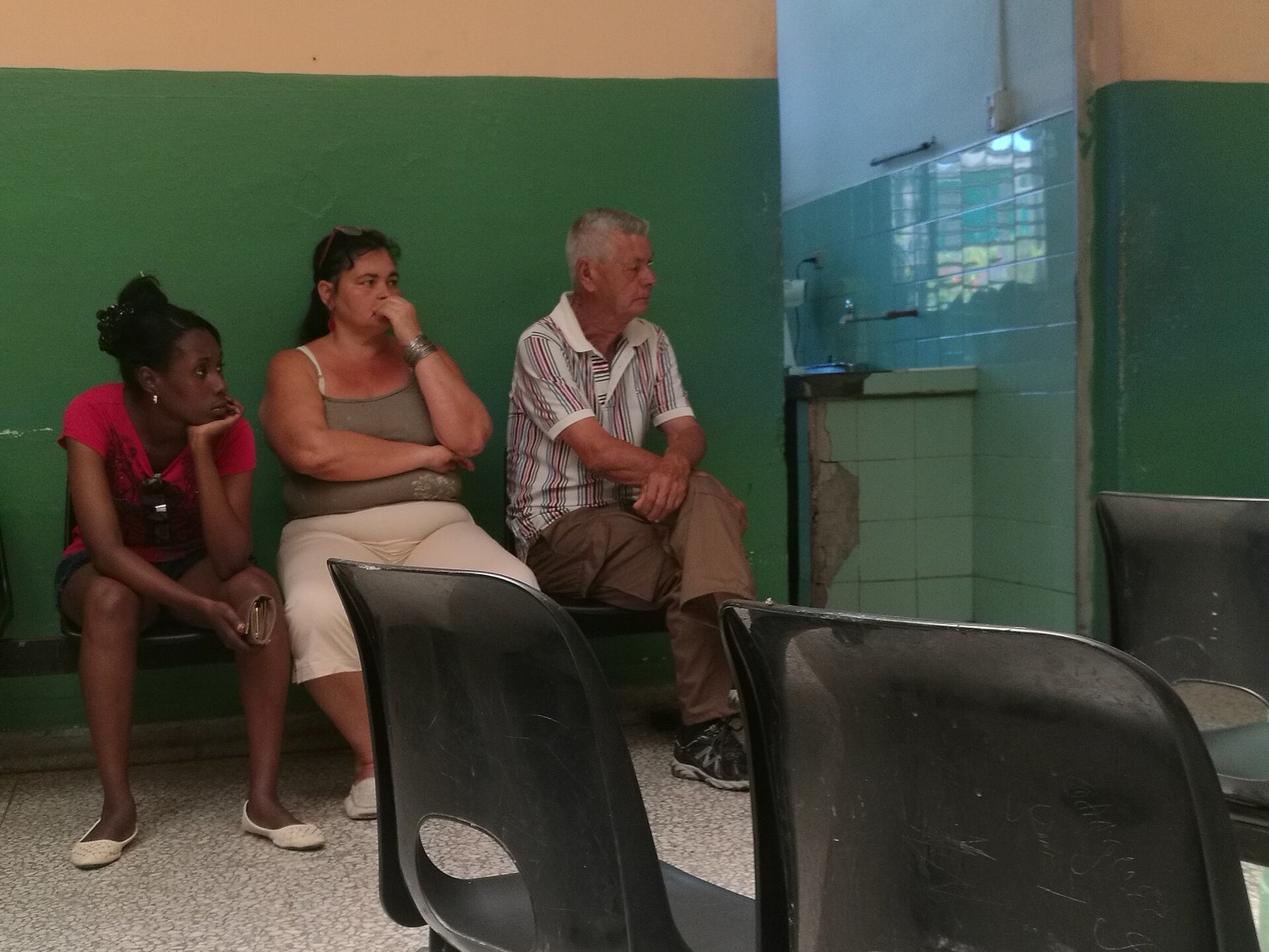 Tres personas esperan sentadas en sillas de plástico negras en una sala de paredes verdes con azulejos rotos.