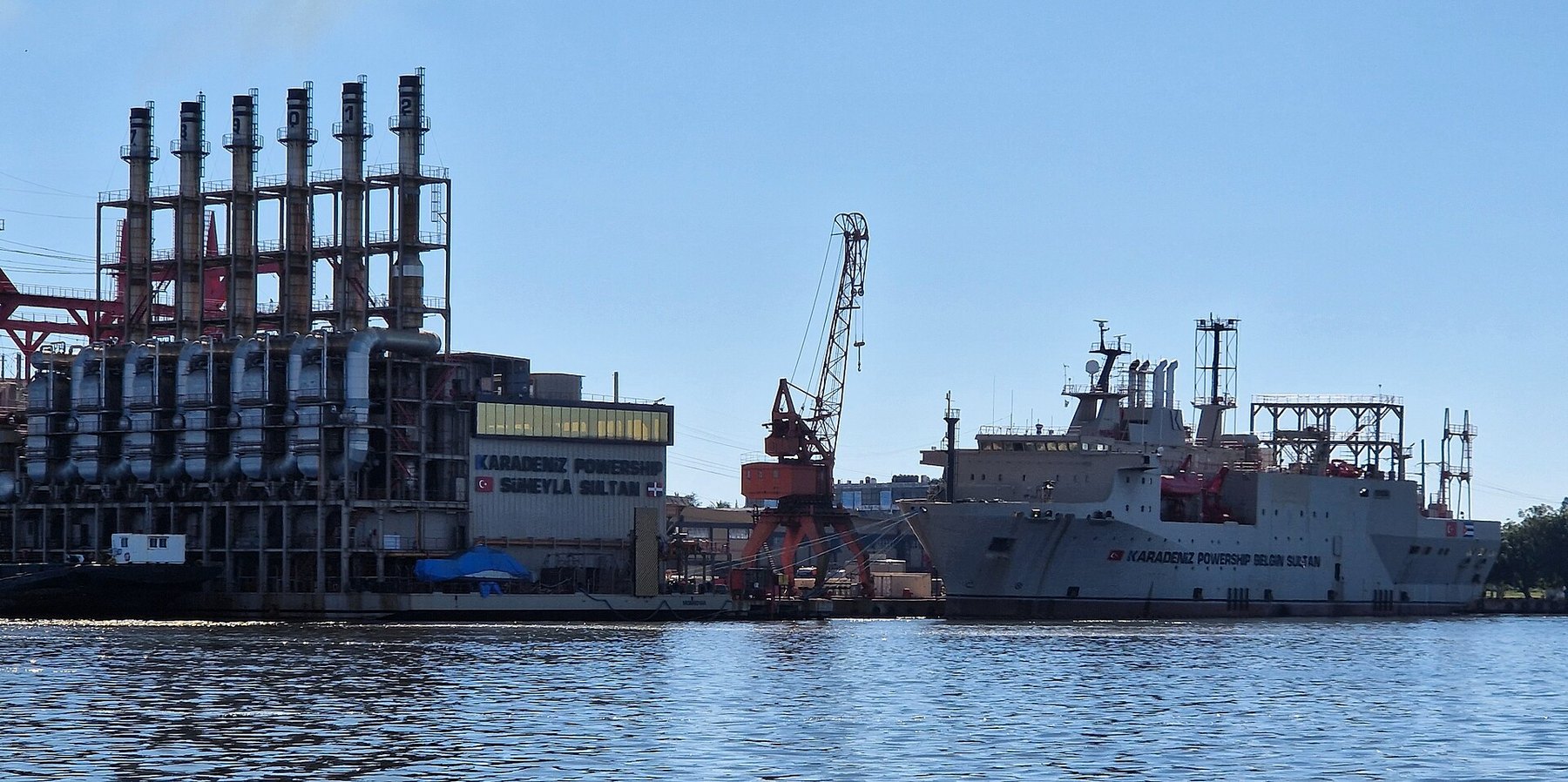 Dos grandes barcos grises con chimeneas y estructuras industriales, amarrados en un muelle con una grúa naranja; en el casco se lee «Karadeniz Powership».